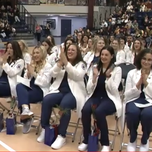 Group of nurses in their navy blue scrubs and white jackets, applauding as they sit in the front row at their pinning ceremony.