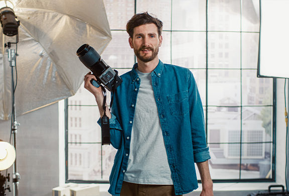 man holding camera with light lamp near him. He has dark hair and beard. He is wearing a blue unbuttoned down shirt with a white t-shirt underneath.