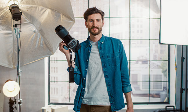 man holding camera with light lamp near him. He has dark hair and beard. He is wearing a blue unbuttoned down shirt with a white t-shirt underneath.