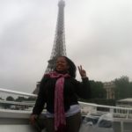 Celina a girl with a red scarf dark skin, and hair, holding her fingers up in the peace sign, with the Paris Eiffel tower behind her.