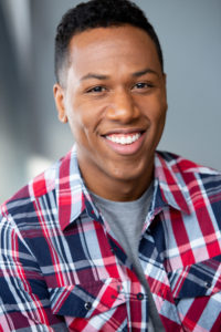 Headshot of Motivational Speaker Aaron K. Wilson. He is wearig a red white and blue plaid shirt with a grey t-shirt showing through. He has a bif smile on his face.