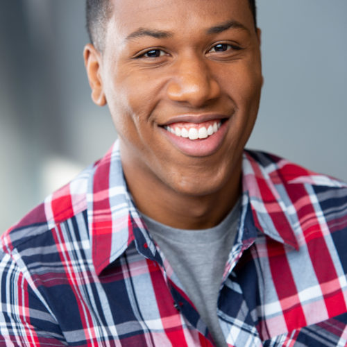 Headshot of Motivational Speaker Aaron K. Wilson. He is wearig a red white and blue plaid shirt with a grey t-shirt showing through. He has a bif smile on his face.