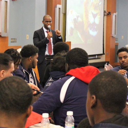 Dark skin male wearing a suit and tie, standing in front of a large audience of dark skin males speaking in a microphone.