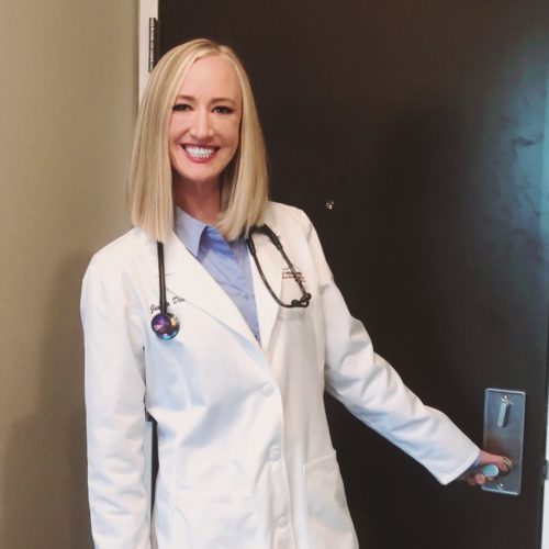 Jessica in doctor's coat and stethiscope around her neck. Blond hair , blue shirt, smiling and about to pull open a door.