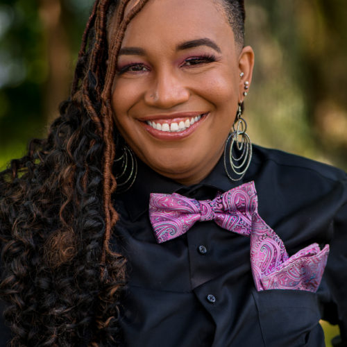 Big smile on a woman with long braided hair wearing a purple bowtie and pocket scarf.