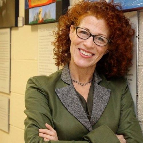 Laura McCullough Brookdale's English Professor Smiling, arms folded, curly red hair and glasses, wearing a green blazer.