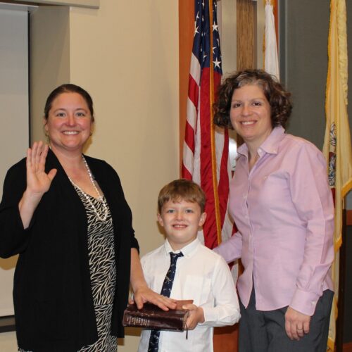 One woman wearing a dress with hand swearing in with young boy wearing a tie, holding the bible. and another woman on the other side of him wearing pants. All smiling.