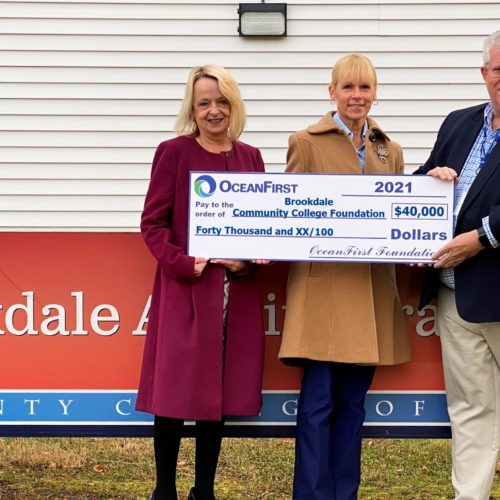 two women wearing coats and one man wearing a suit jacket, holding a large check in front of the Brookdale Community College Administration Building sign outside