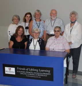 A group of "Friends of Lifelong Learning" Five standing and 3 sitting behind a table with a blue tablecloth and sign Friends of Lifelong Learning