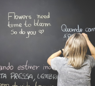 woman with blond hair writing on the chalk board translating a phrase Flowers need time to bloom so do you in another language