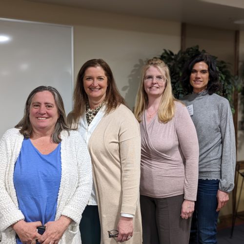 Four women standing and smiling with casual clothes on.