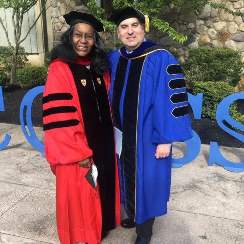 A black woman in red Regalia and a white man in royal blue Regalia standing in front of a Success sculpture.