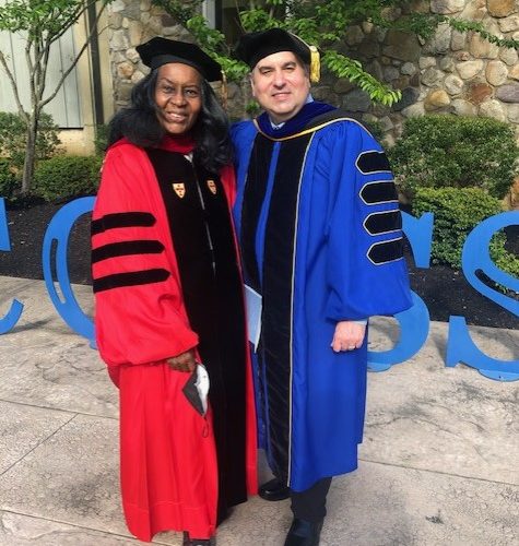 A black woman in red Regalia and a white man in royal blue Regalia standing in front of a Success sculpture.