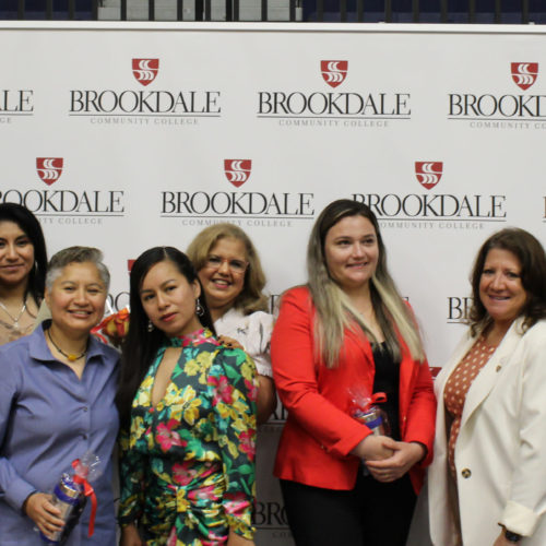nine women and a man dressed nicely and smiling with awards in their hands and a backdrop of Brookdale Community College logos behind them