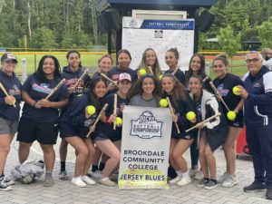 group of women softball team members gathered with the tournament brackets behind them. They are holding bats and yellow softballs. All wearing navy blue shorts and t-shirts