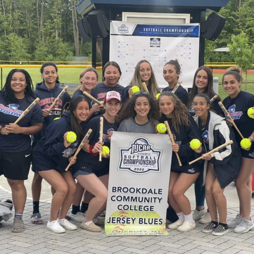 group of women softball team members gathered with the tournament brackets behind them. They are holding bats and yellow softballs. All wearing navy blue shorts and t-shirts