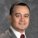 Man with dark hair smiling wearing a suit and white shirt and dark red tie.