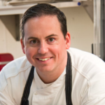 male with short brown hair wearing an apron and white chef jacket