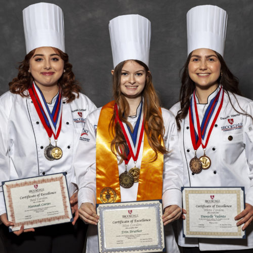 three girls wearing chef jackets and hats with medals around their neck and certificates in their hands
