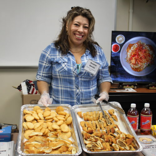 Hispanic woman holding trays of sample food at her expo booth.