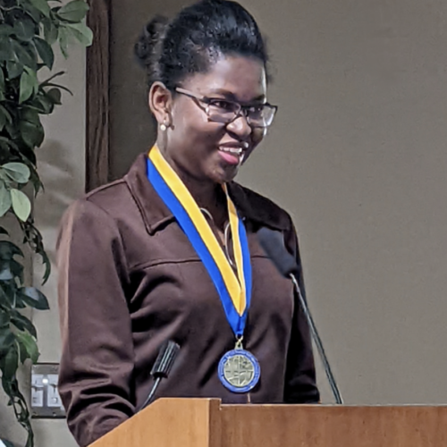 Dark skinned woman wearing a brown shirt and has a medal hanging around her neck.