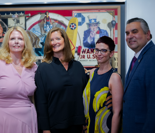 Three women wearing dresses and a man wearing a suit smiling in front of a very large colorful painting.