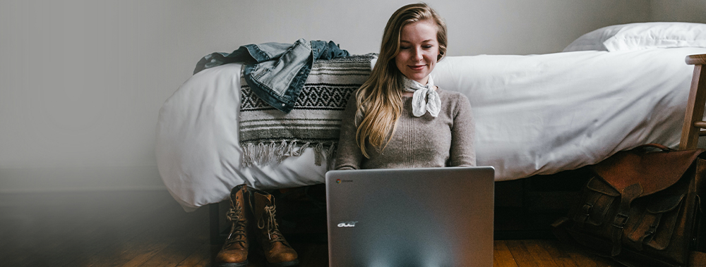 A woman sitting on the floor next to her bed with a computer on her lap.