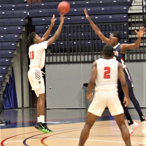 Three basketball players. One shooting the ball, one with hands up to try to block the ball and one looking on.