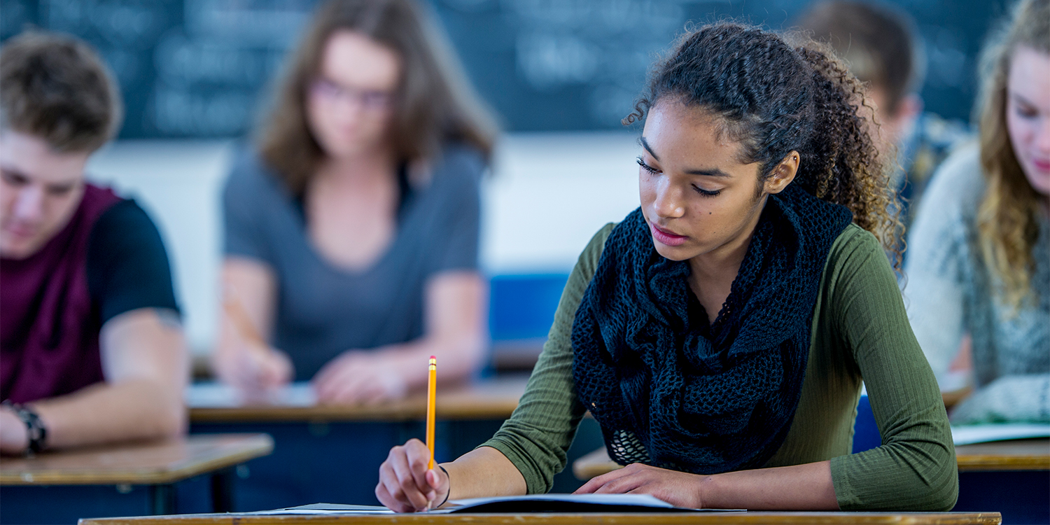 Woman with dark skin at a desk with pencil in her hand writing on an open notebook. Other students in the background in similar activity.