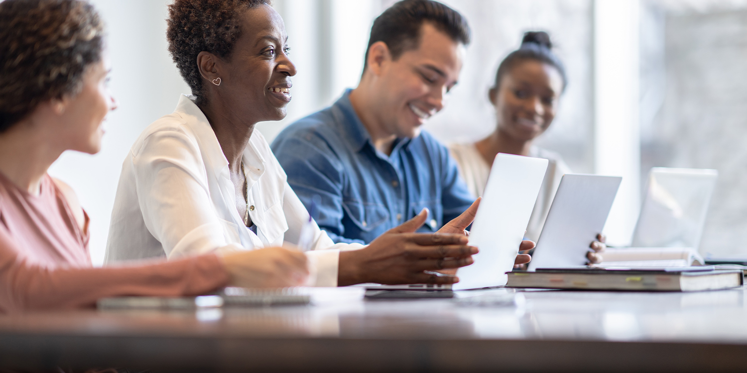 Older dark skinned woman smiling with her laptop open and four other students on either side of her also smiling. One male with a blue shirt on to her left and two women.