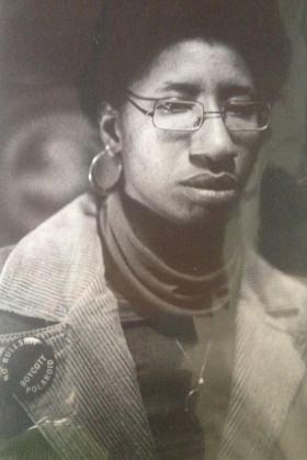 Photograph of a dark skinned woman wearing glasses, hoop earrings, turtleneck and corduroy jacket that has a pin on the lapel.