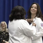 Two nurses in their white jackets. One is pinning the other with two woman in the background smiling.