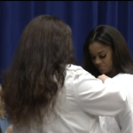 Two nurses. One pinning the other. Woman smiling in the backgroud.