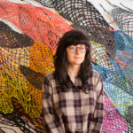 Woman with long dark hair and bangs, wearing glasses and a plaid shirt with her colorful art work of woven paper sculptures behind her.