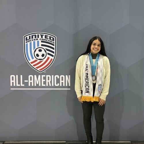 All American shield with soccer ball in the middle. Woman with long dark hair wearing a white sweater and dark pants and a sash around her neck that says All American.
