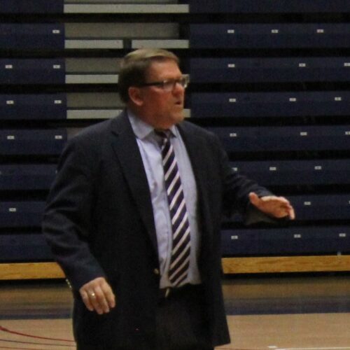 Man in dark suit and striped tie with light blue shirt, wearing glasses in the gymnasium.