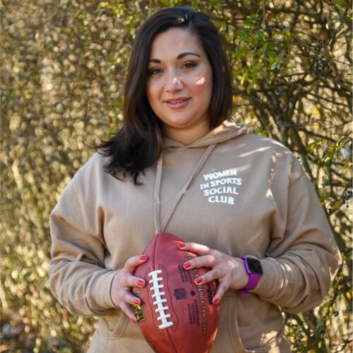 Woman with dark long hair wearing a beige sweatshirt and holding a football.