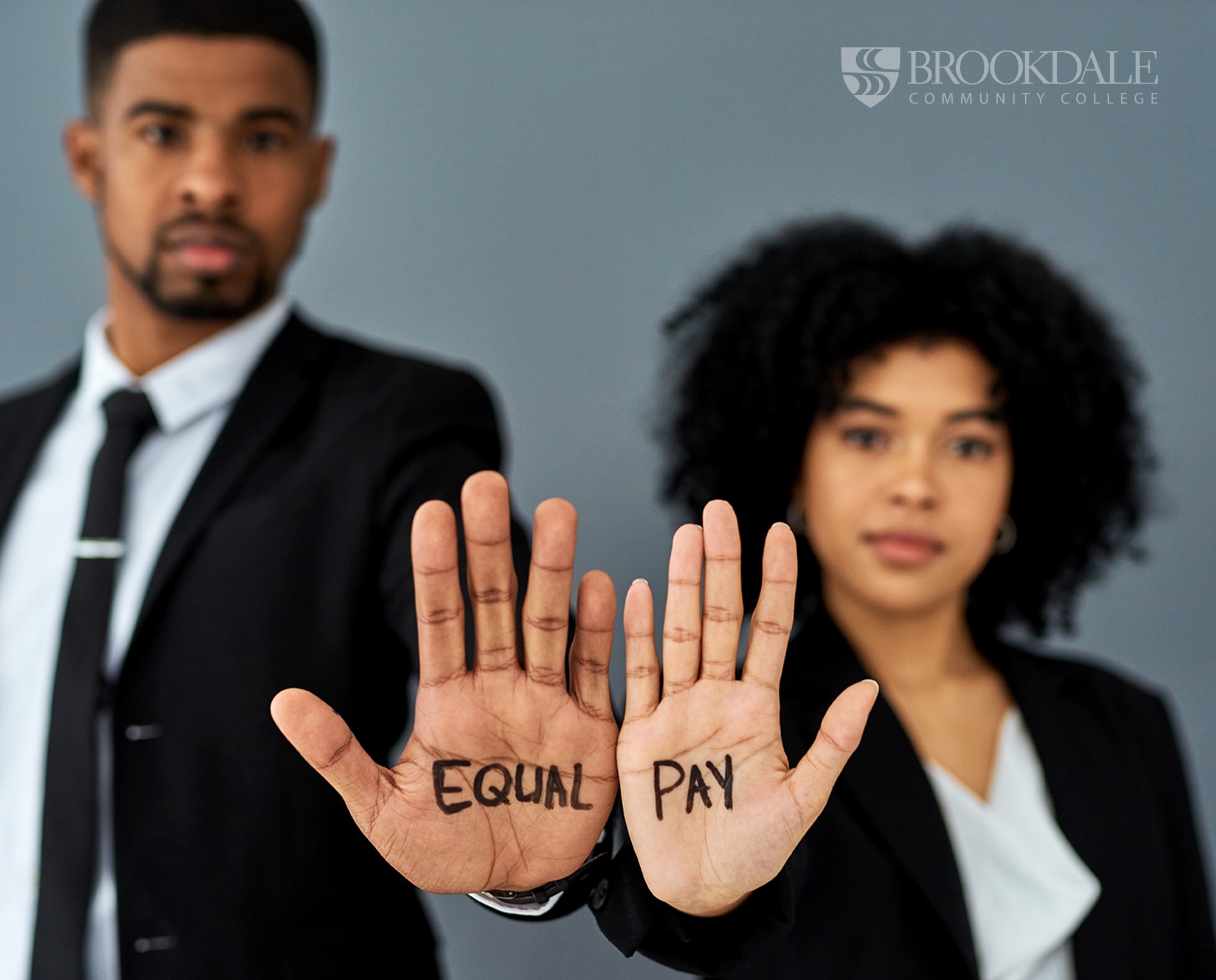 Two persons of color one male and one female holding their hands up with the male palm reading Equal and the female palm reading Pay. Both professionally dressed.