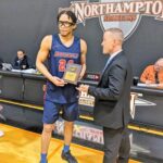Male basketball player wearing navy blue with red number and letters that read Brookdale number 24. He is wearing glasses and ankle socks and black sneakers. He is holding his trophy and a man in a suit is presenting it to him.