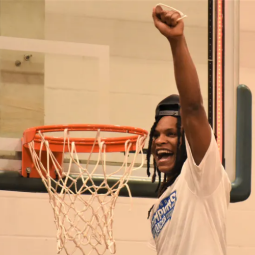 Dark skinned man with a big smile on his face, raising his hand in the air with his face next to the basketball hoop after cutting a piece of the rope from it.