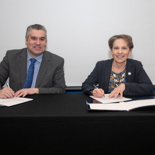 Man and woman wearing suits and signing papers on a black tabletop.