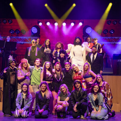Group of 20 men and women on stage with lights shining down on them. All actors in the Rock of Ages at the Performing Arts Center in Lincroft, NJ.