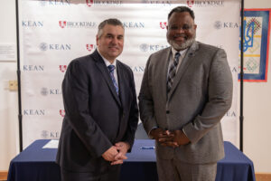 Two men dressed in suit and ties smiling. One man is light skin and wearing dark grey suit and blue tie and the other is dark skin man wearing a tan suit.