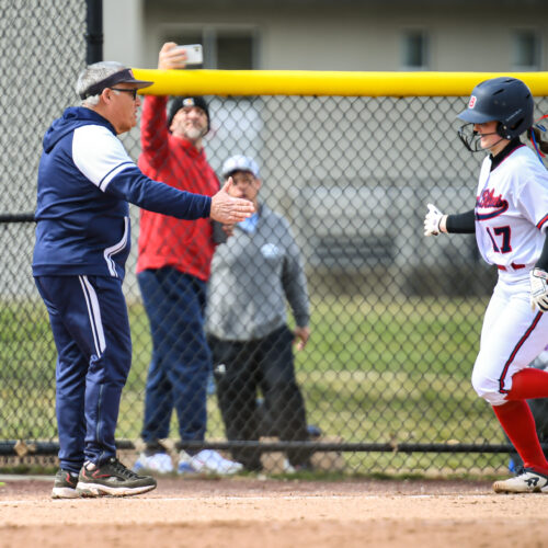 Coach wearing navy blue sweat pants and jacket high fives softball runner.