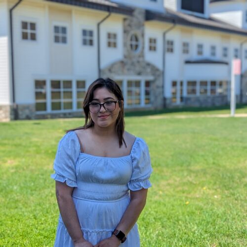 Woman with dark hair and wearing a white dress smiling in front of college building.