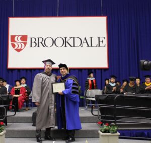 Honorary Degree Douglas Quinn receiving award from President Dr. David M. Stout on stage at Commencement ceremony.