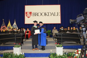 Sean P. O'Halloran receiving Outstanding Alum Award on stage at Commencement ceremony wearing cap and gown along with President Stout presenting the award.
