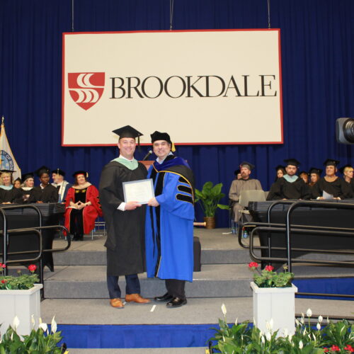 Sean P. O'Halloran receiving Outstanding Alumni Award on stage at Commencement ceremony wearing cap and gown along with President Stout presenting the award.