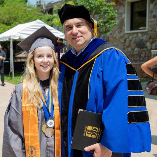 Woman with long blond hair in her cap and gown and gold honor society regalia with Dr. Stout also in his royal blue regalia after commencement ceremony.