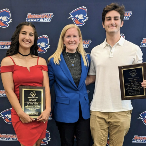 A male and female athlete on either side of the Athletic Director. They are holding awards in their hands.